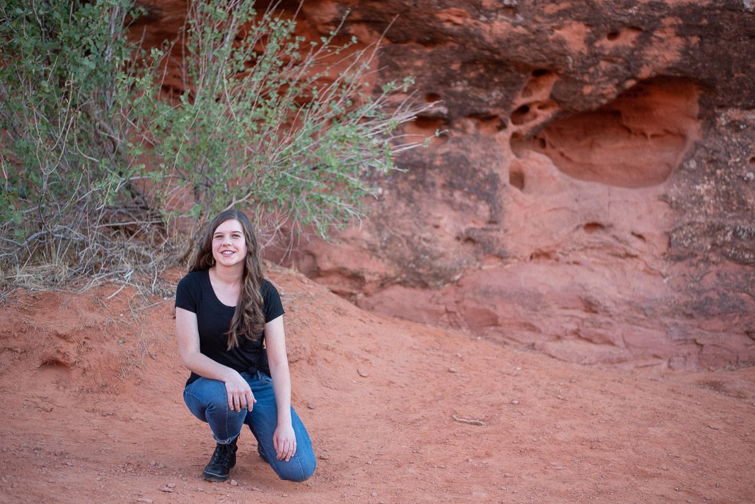Teenager kneeling down in a red rock area with a bush in the background